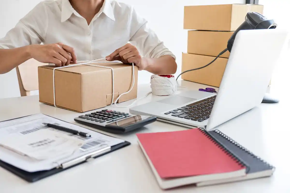 Person packing a box for shipping, with a laptop, calculator, and shipping labels on the table, representing e-commerce platform order fulfillment.